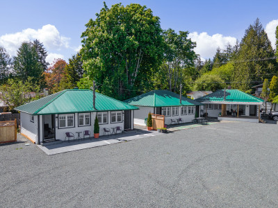 Aerial view of three beach cabins with green roofs, set in a spacious gravel area at ROAM Beach Resort. The cabins are surrounded by lush trees under a clear blue sky. The area is open and inviting.