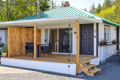 A beach cabin at ROAM, labelled Cabin 5, with a green roof and a dark grey door. It features a wooden porch with a small table and two chairs, partially enclosed by a wooden privacy screen.