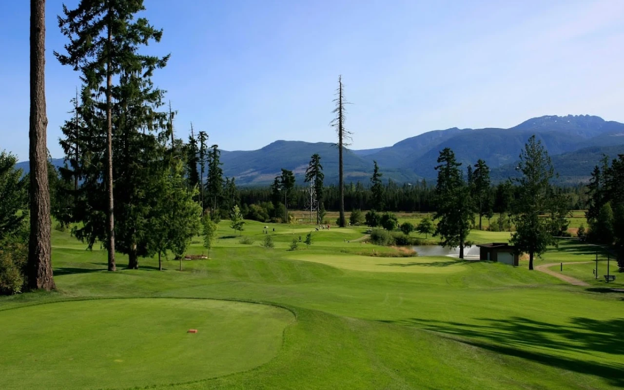 A scenic view of Arrowsmith Golf and Country Club, showcasing a lush green golf course surrounded by tall trees and rolling hills. In the background, mountains rise against a clear blue sky, creating a picturesque setting.