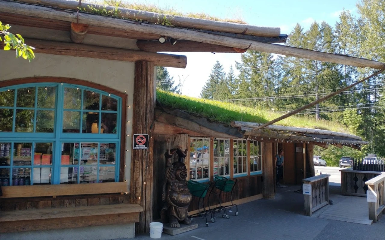 A charming building at Coombs Country Farms featuring a unique green roof adorned with grass and plants. The structure has rustic wooden beams and large windows framed in blue, creating a welcoming atmosphere.