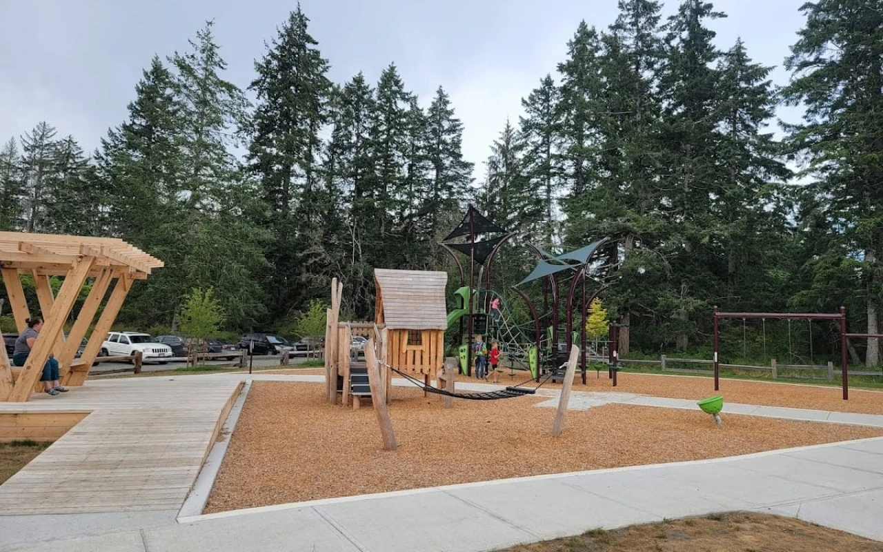 A modern playground set within Dunsmuir Community Park, featuring a wooden playhouse and a large climbing structure adorned with green and black elements.