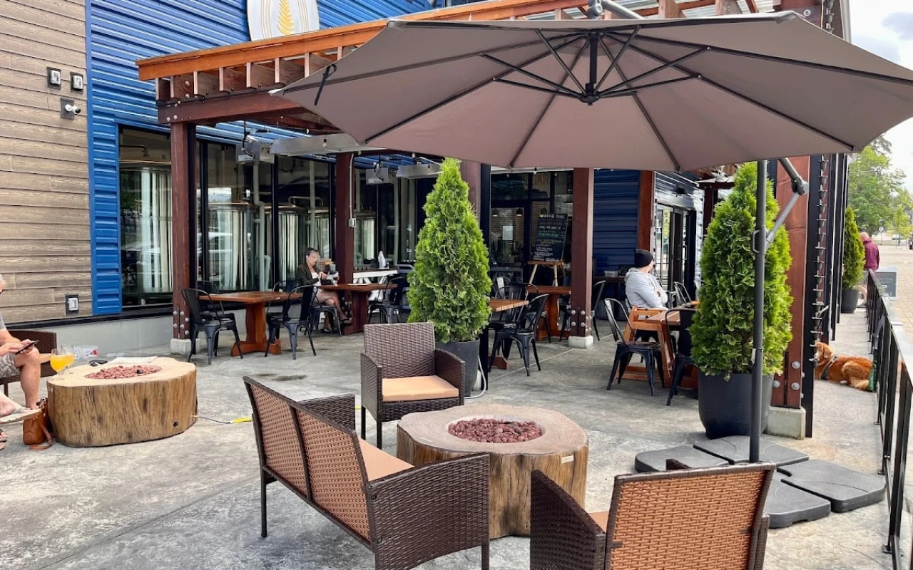 An inviting outdoor seating area of a brewery, featuring wicker chairs and tables arranged around a wooden fire pit. Large umbrellas provide shade, and potted greenery adds a touch of nature.