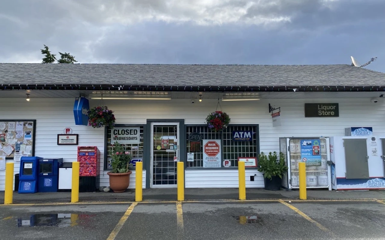 A quaint store front with a welcoming entrance featuring a sign that reads "OPEN". The building has white siding and a sloped roof, with hanging flower baskets adding charm. An ATM and a sign for a liquor store are visible to the right.