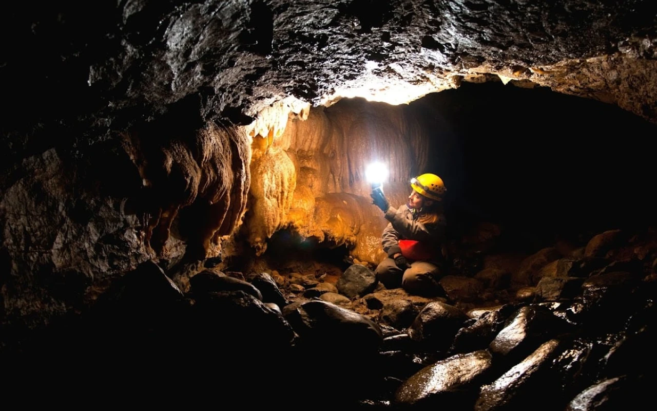 A person wearing a yellow helmet and holding a bright light is exploring a cave. The cave walls are rugged and uneven, illuminated by the light, revealing intricate rock formations. The ground is covered with wet, smooth stones, and the atmosphere is dark and mysterious.