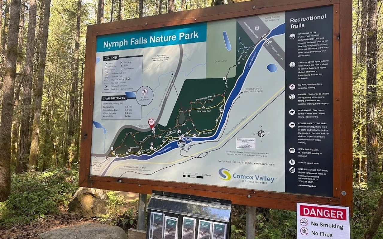 A detailed information board for Nymph Falls Nature Park, displaying a map of the park's trails and key features. Surrounding the sign are tall trees and lush vegetation, creating a natural and inviting atmosphere.