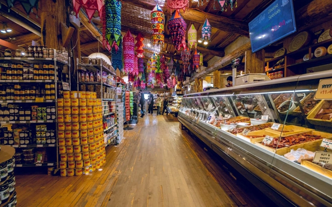 An interior view of a vibrant market featuring an array of colourful hanging decorations. The wooden floors add a rustic charm, while shelves stocked with various canned goods line the sides.