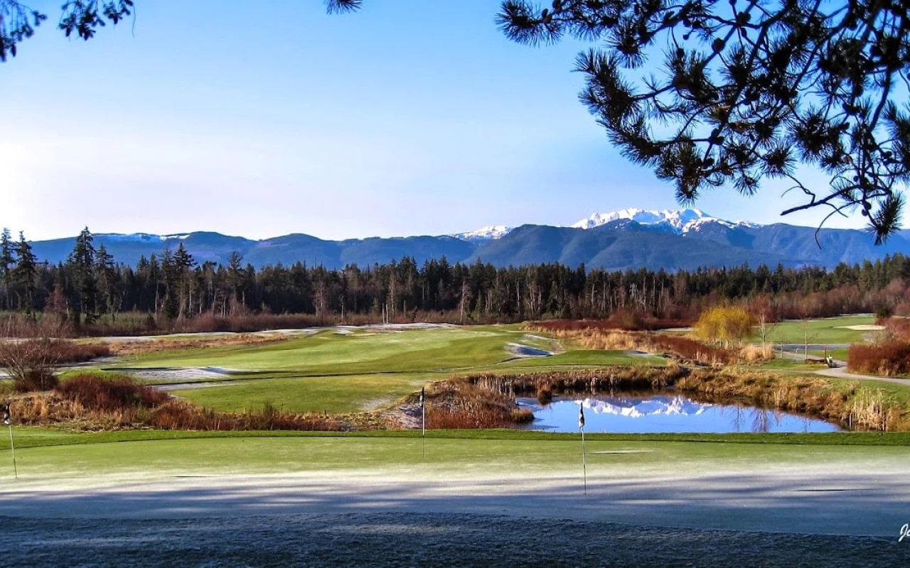 A scenic view of a golf course featuring lush green fairways and a small pond reflecting the clear blue sky. In the background, a line of evergreen trees stands before a range of snow-capped mountains.