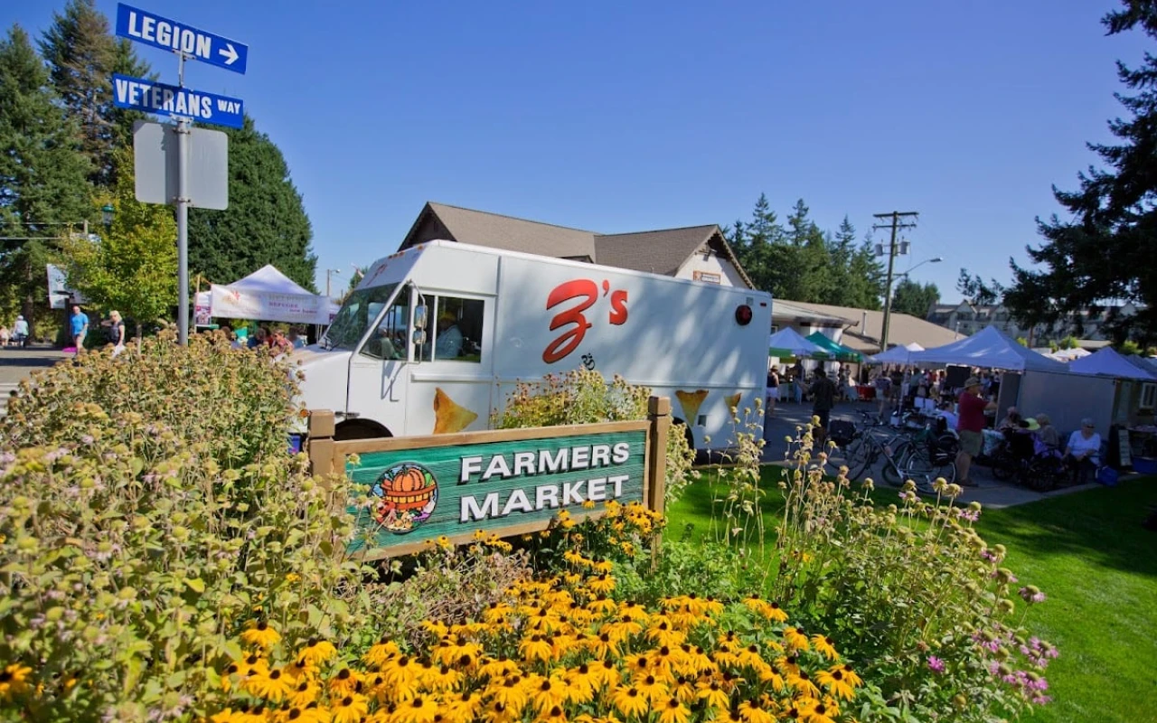 A vibrant scene at a farmers' market, featuring a sign that reads "FARMERS MARKET" in front of a food truck. Bright yellow flowers and greenery surround the area, adding colour to the setting. In the background, several vendor tents are visible.
