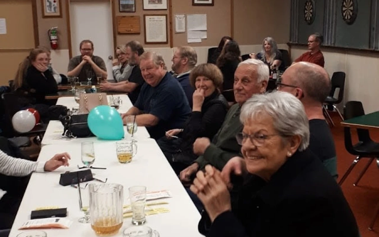 A group of people seated around a long table in a community hall setting. The table is covered with glasses and pitchers, and there are colourful balloons as decorations.