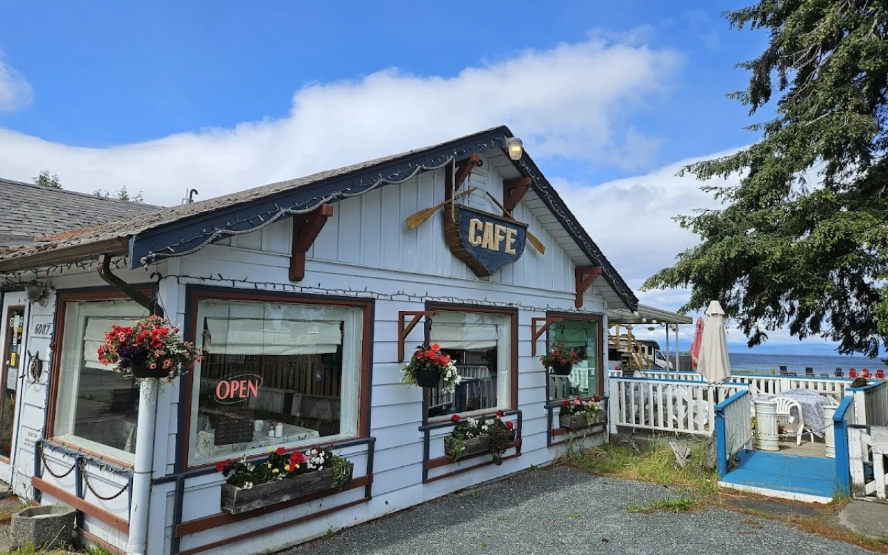 A quaint café with a rustic exterior, featuring white wooden walls and a large "Café" sign shaped like a boat with oars. The windows are adorned with flower boxes filled with colourful blooms.