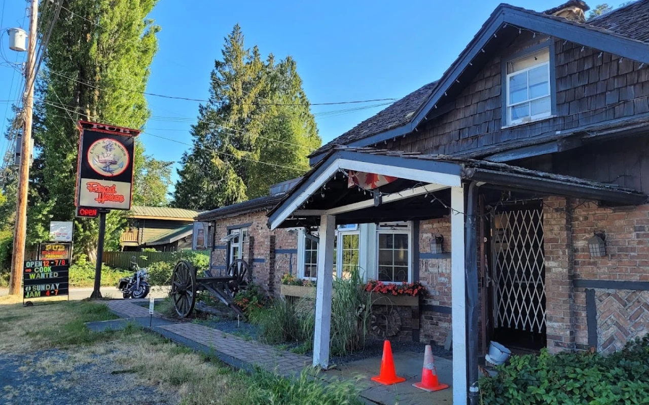 A charming pub with a rustic brick exterior and a wooden shingle roof. A sign with "Pub" and "Road House" hangs near the entrance. Flower boxes beneath the windows add a touch of colour, and an old wagon wheel is displayed as decoration.