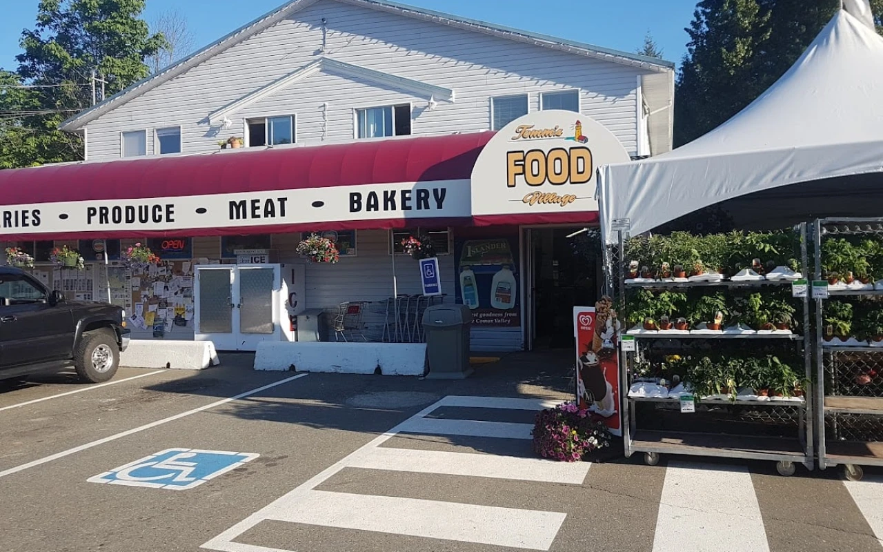 A welcoming food market featuring a large awning. The entrance is adorned with hanging flower baskets, and a sign indicates that the store is open. In front of the market, a black vehicle is parked, while a tent displays potted plants for sale.
