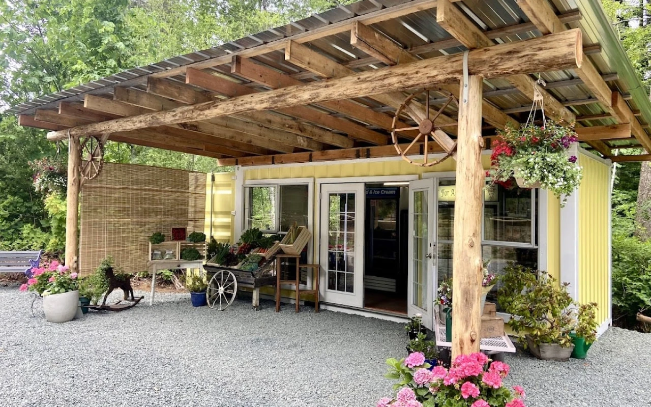 A charming farm shop with a yellow exterior and a wooden overhang supported by rustic beams. The entrance features large glass doors, inviting customers inside.